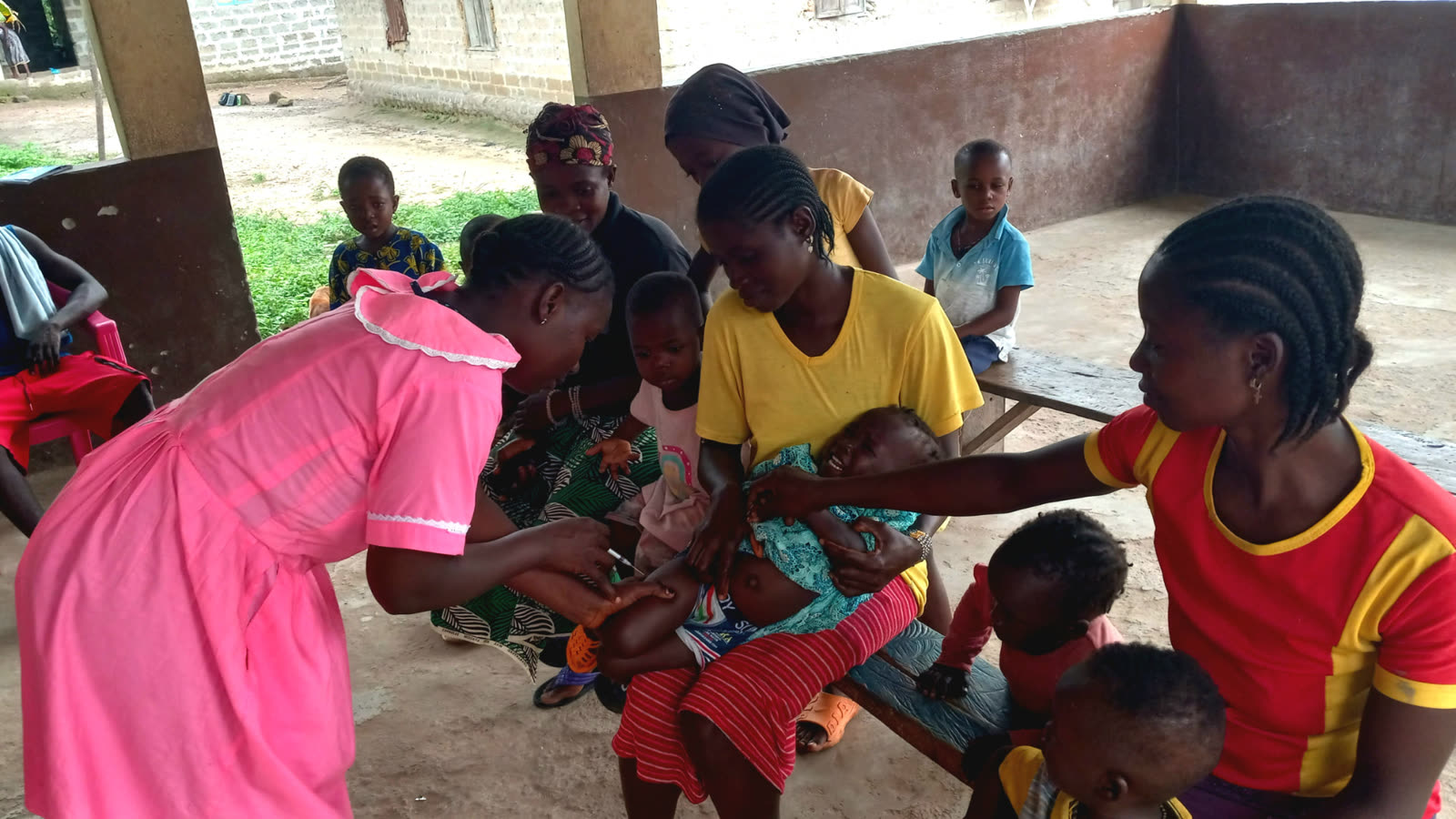 A health worker in pink attending to a young child held on a woman's lap, with other women and children seated nearby