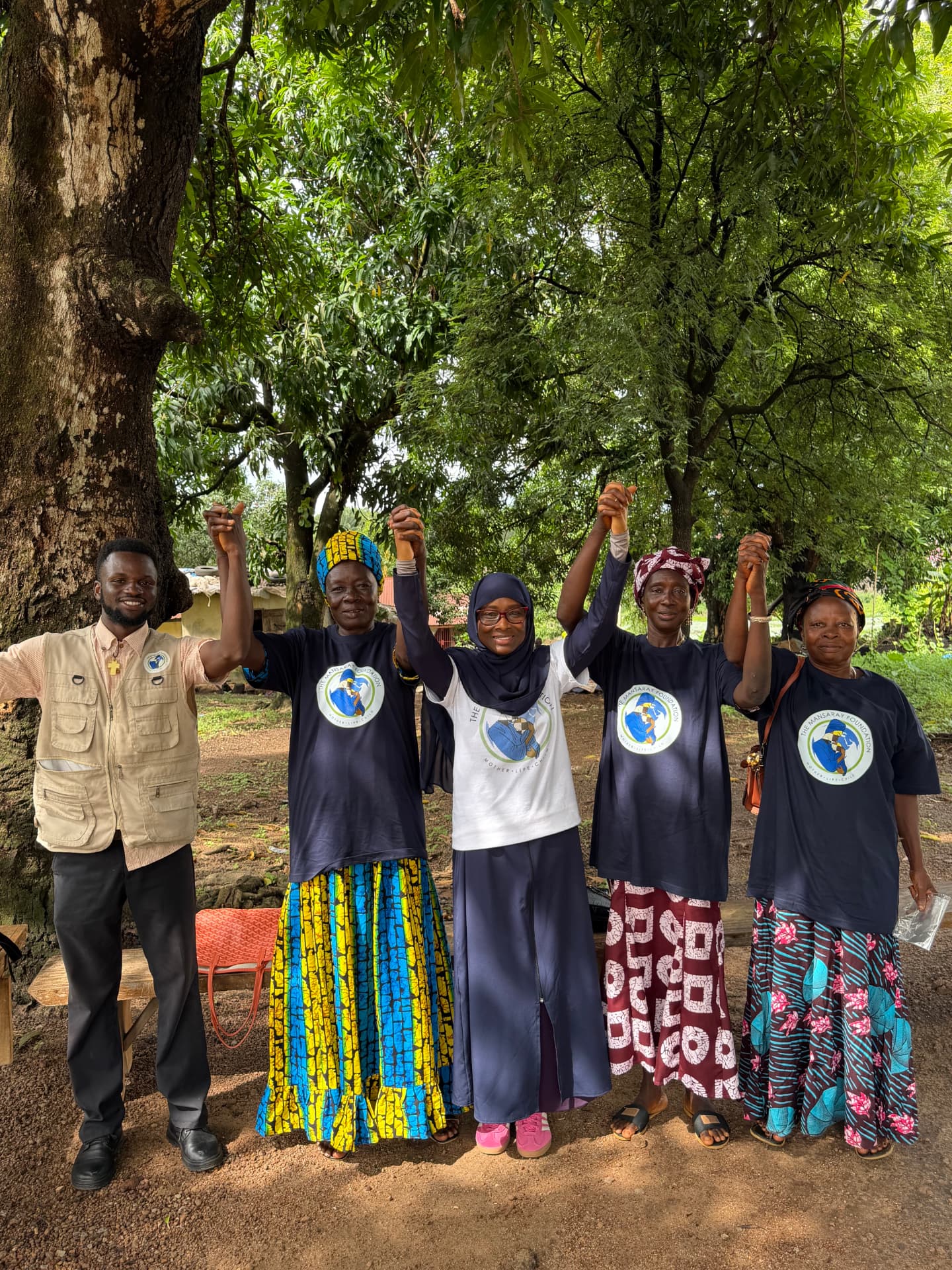 Community partners in TMF shirts standing with raised arms beside a TMF team member beneath a tree