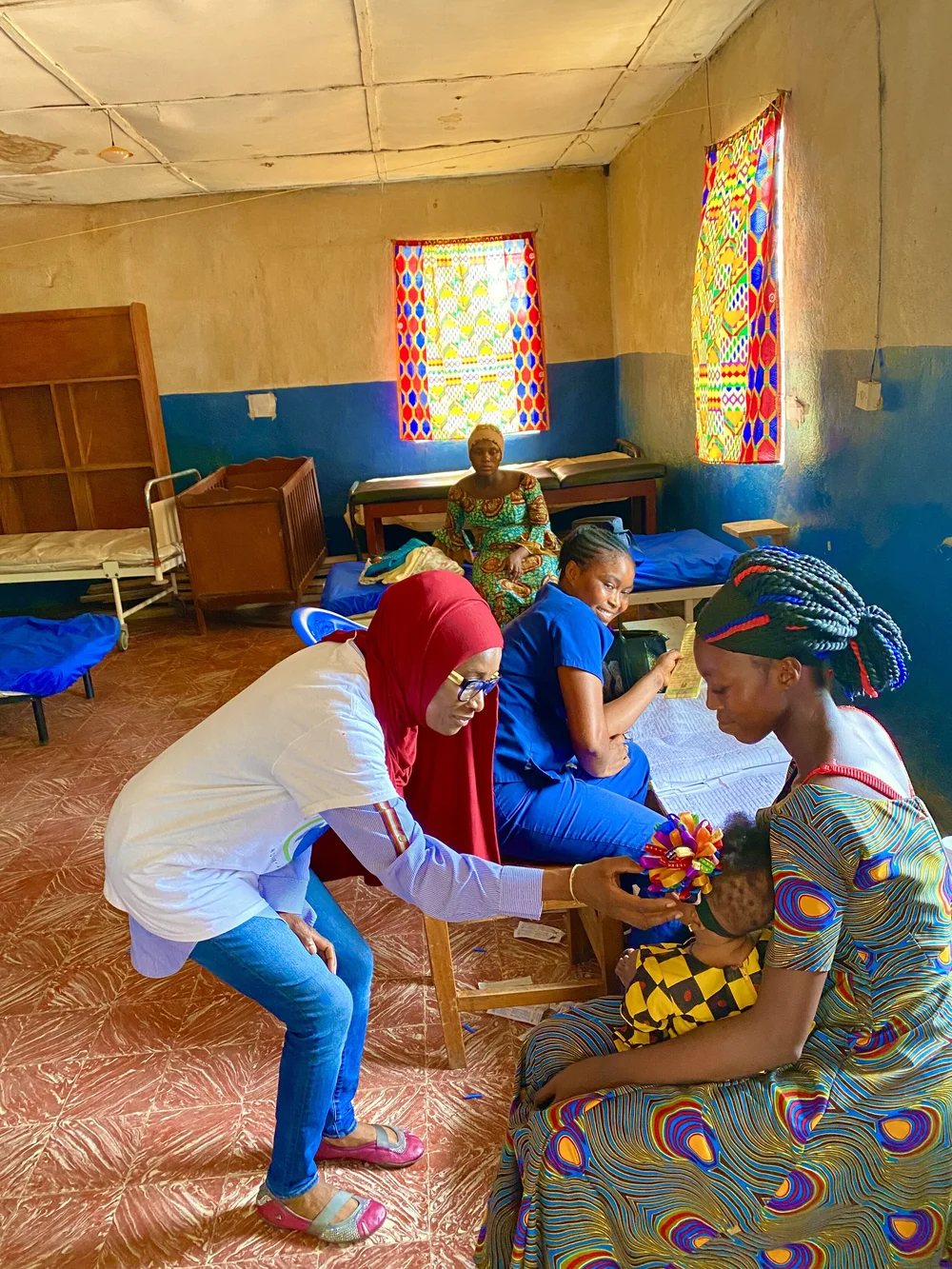 A health worker in a red headscarf leaning toward a young child held by a seated woman inside a clinic room