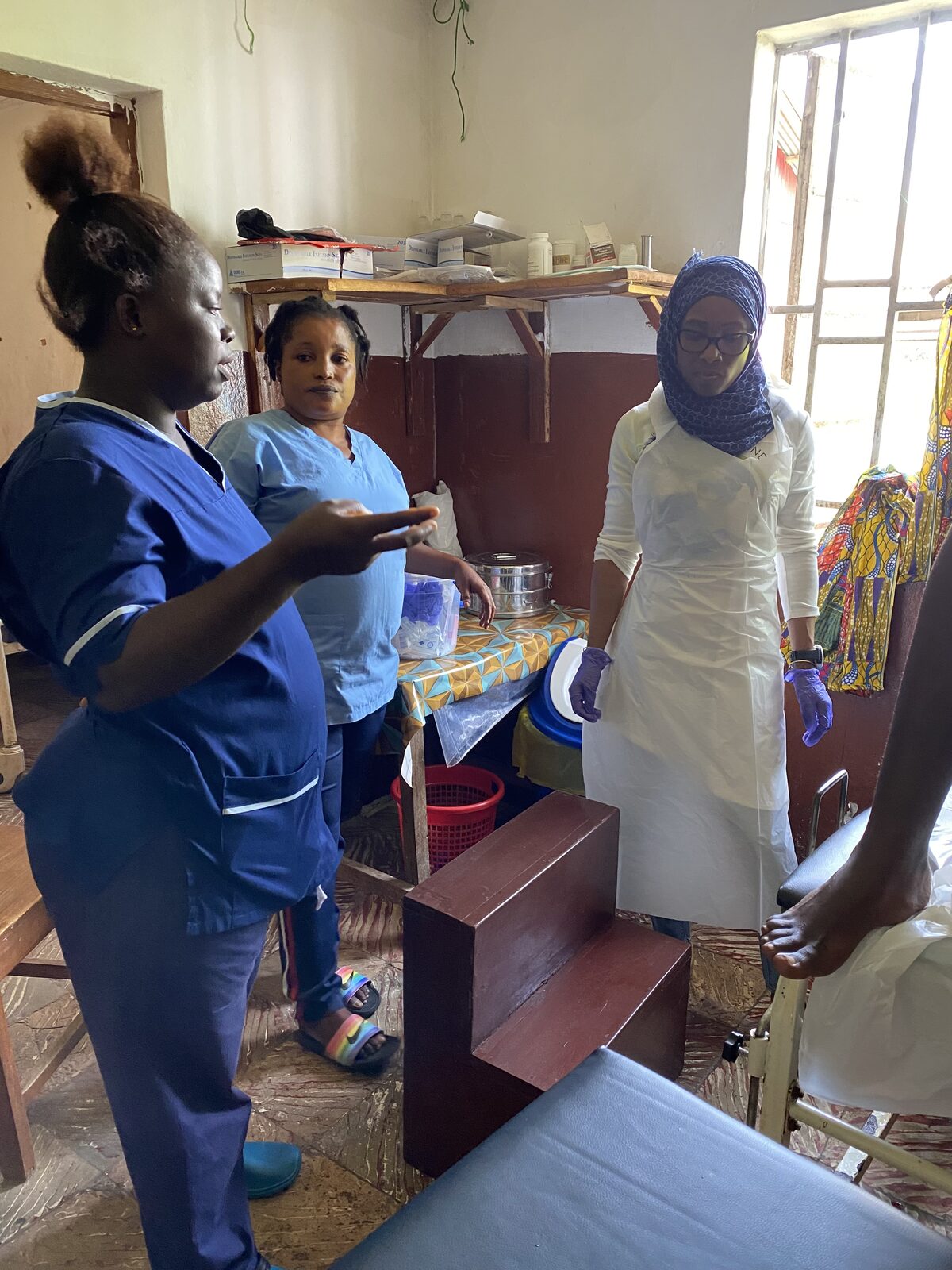 Three health workers in scrubs and protective aprons talking together inside a clinic treatment room