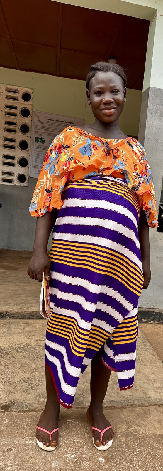 A pregnant woman in an orange top and striped wrap standing outside a health facility