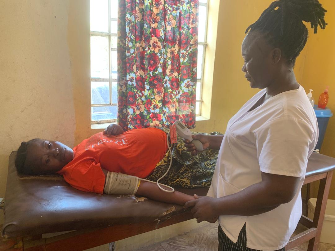 A health worker holding a CRADLE vital signs device while a woman lying on an examination bed wears a blood-pressure cuff