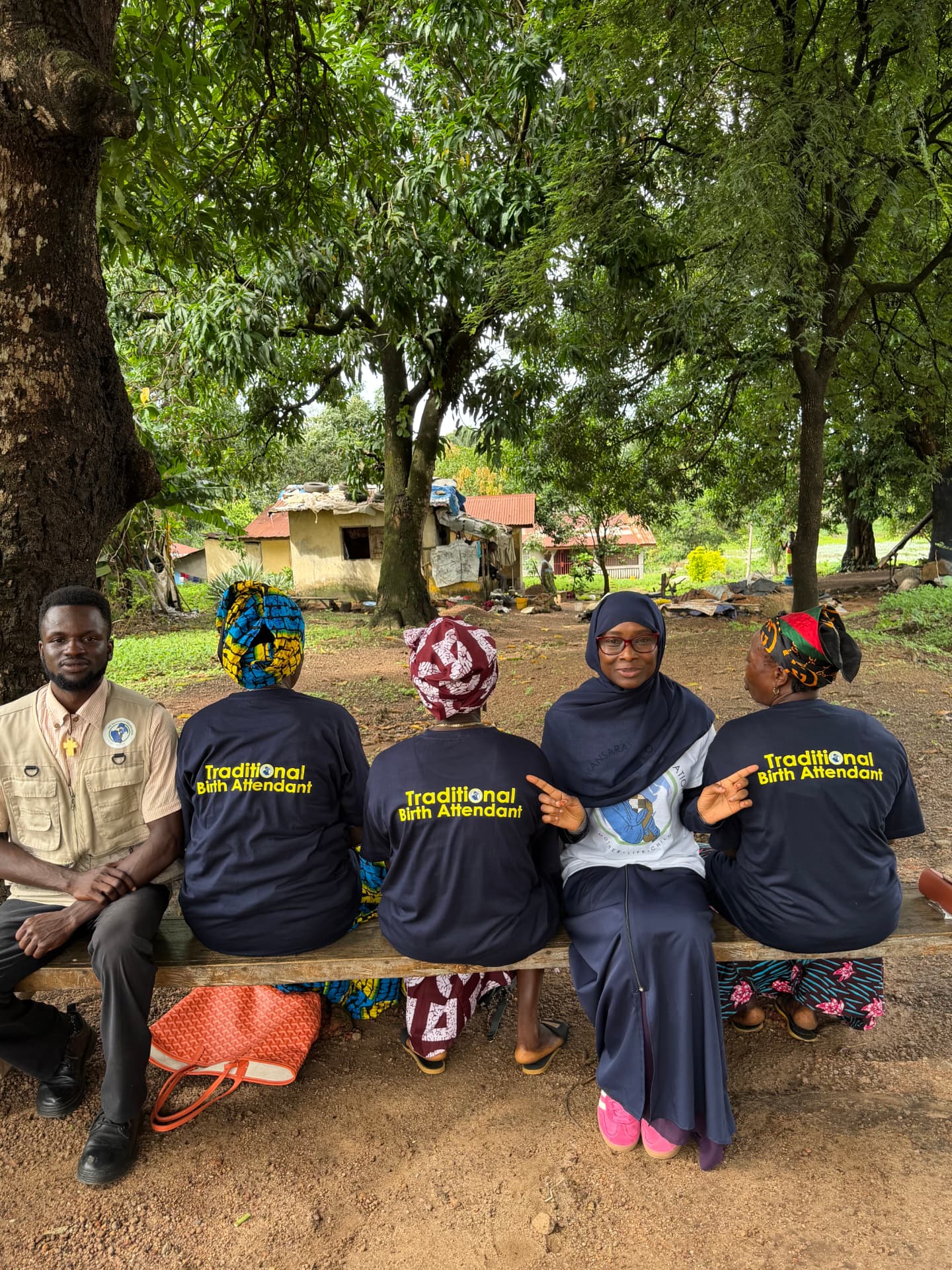 Traditional Birth Attendants in labeled shirts seated with a TMF team member beneath a tree