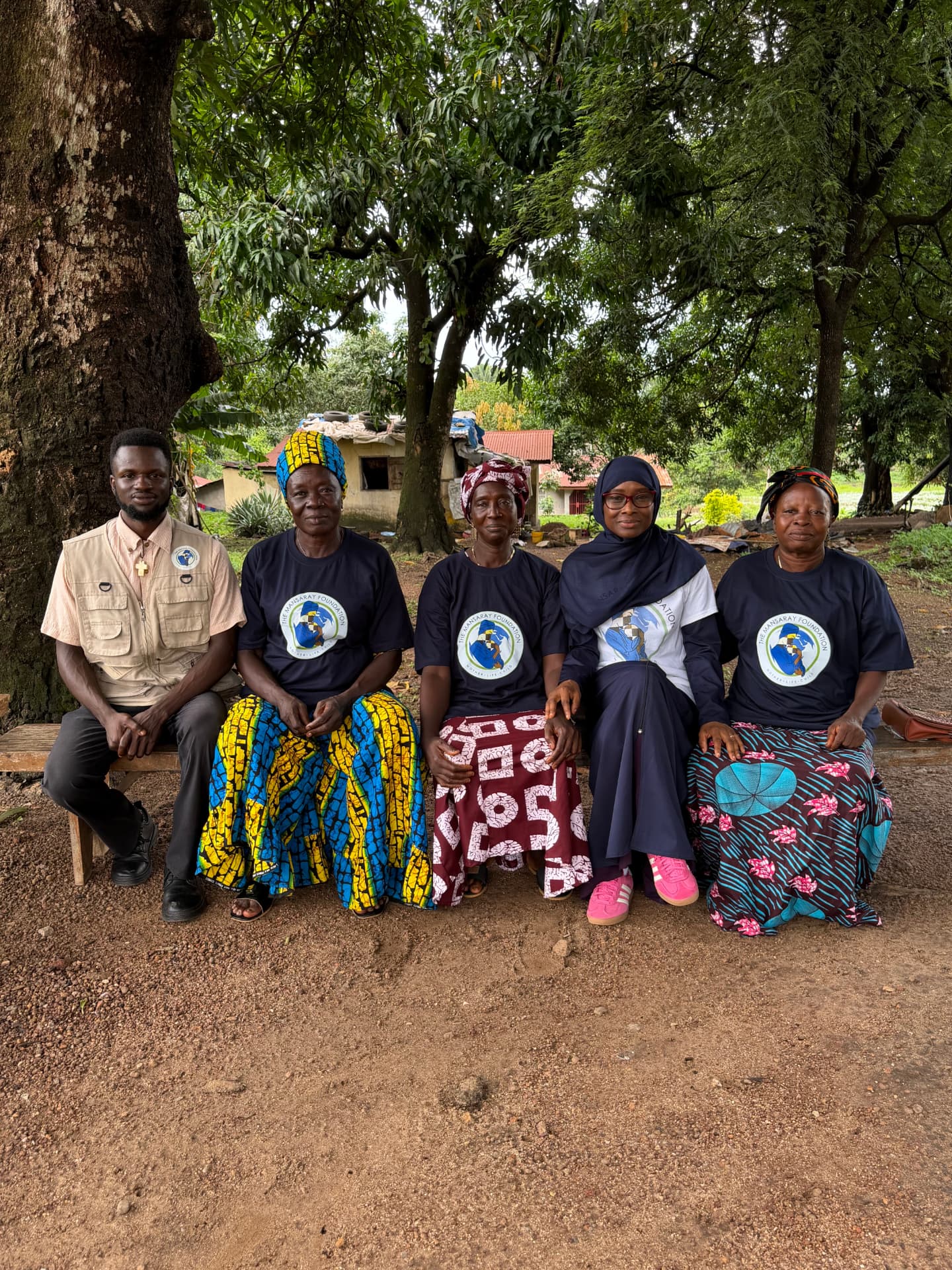 Community women in Mansaray Foundation shirts seated on a bench outdoors with a TMF team member