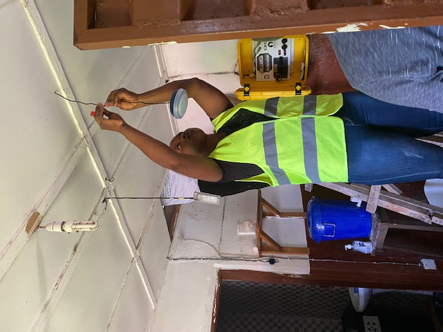 A worker in a high-visibility vest on a ladder wiring a ceiling light beside a wall-mounted control box