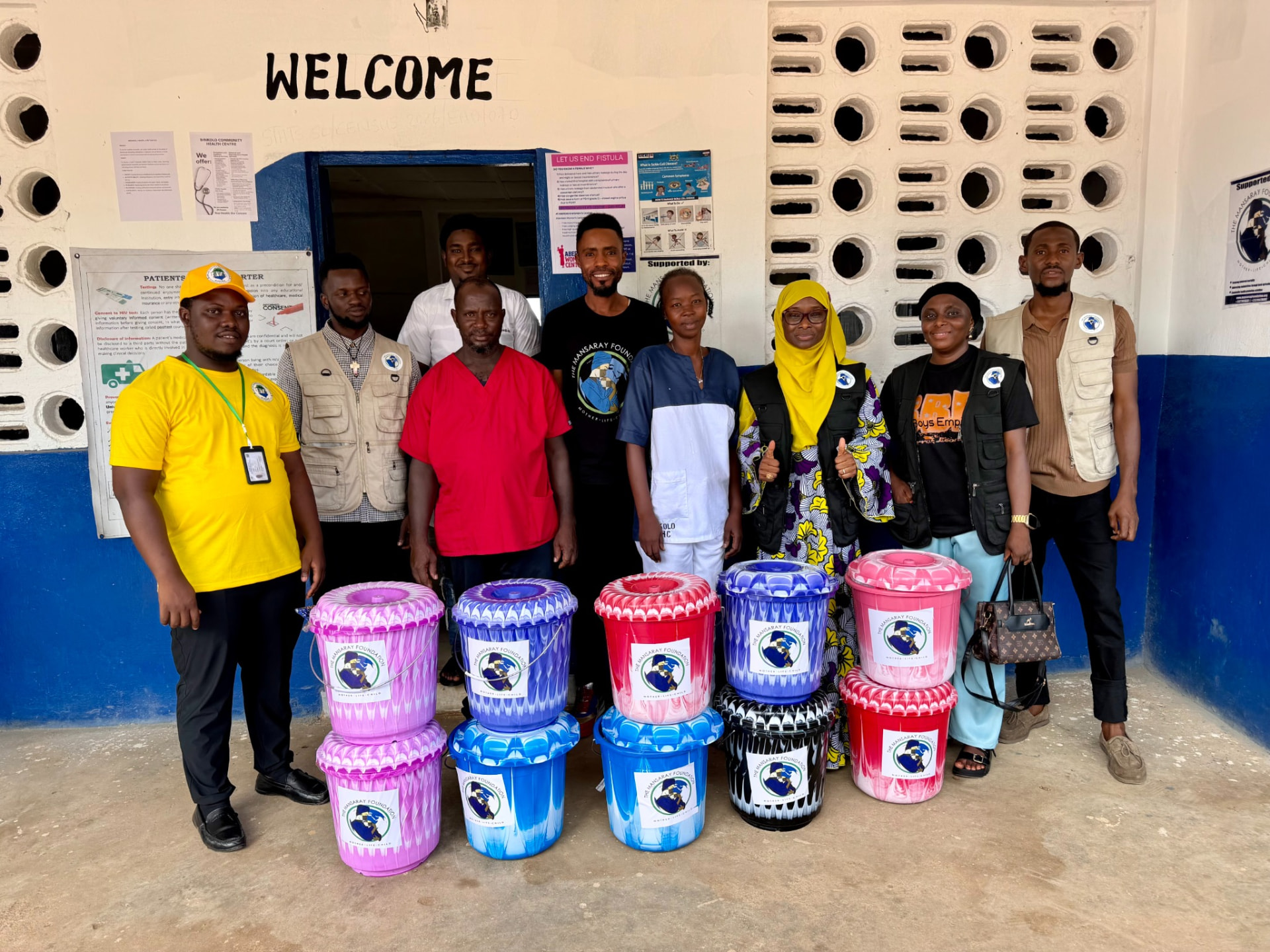 Health workers and TMF team members standing at a clinic entrance beside stacked birthing buckets