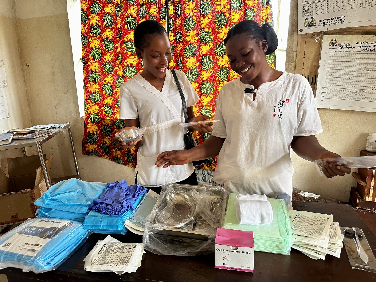 Two health workers in white smiling over medical supplies laid out on a table, including gowns, gloves, needles, and instrument trays