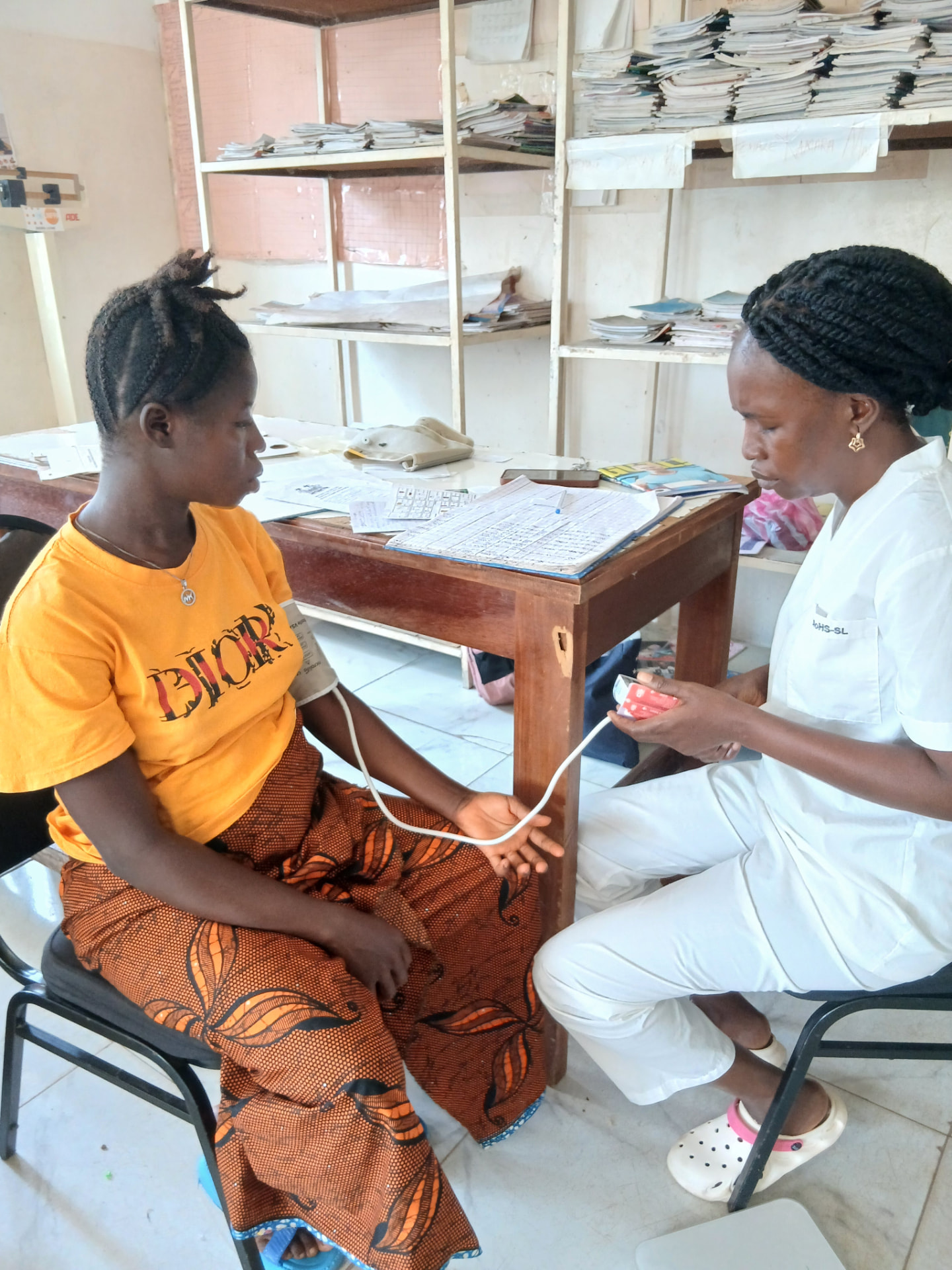 A health worker in white using a CRADLE vital signs device with a blood-pressure cuff on a seated woman's arm