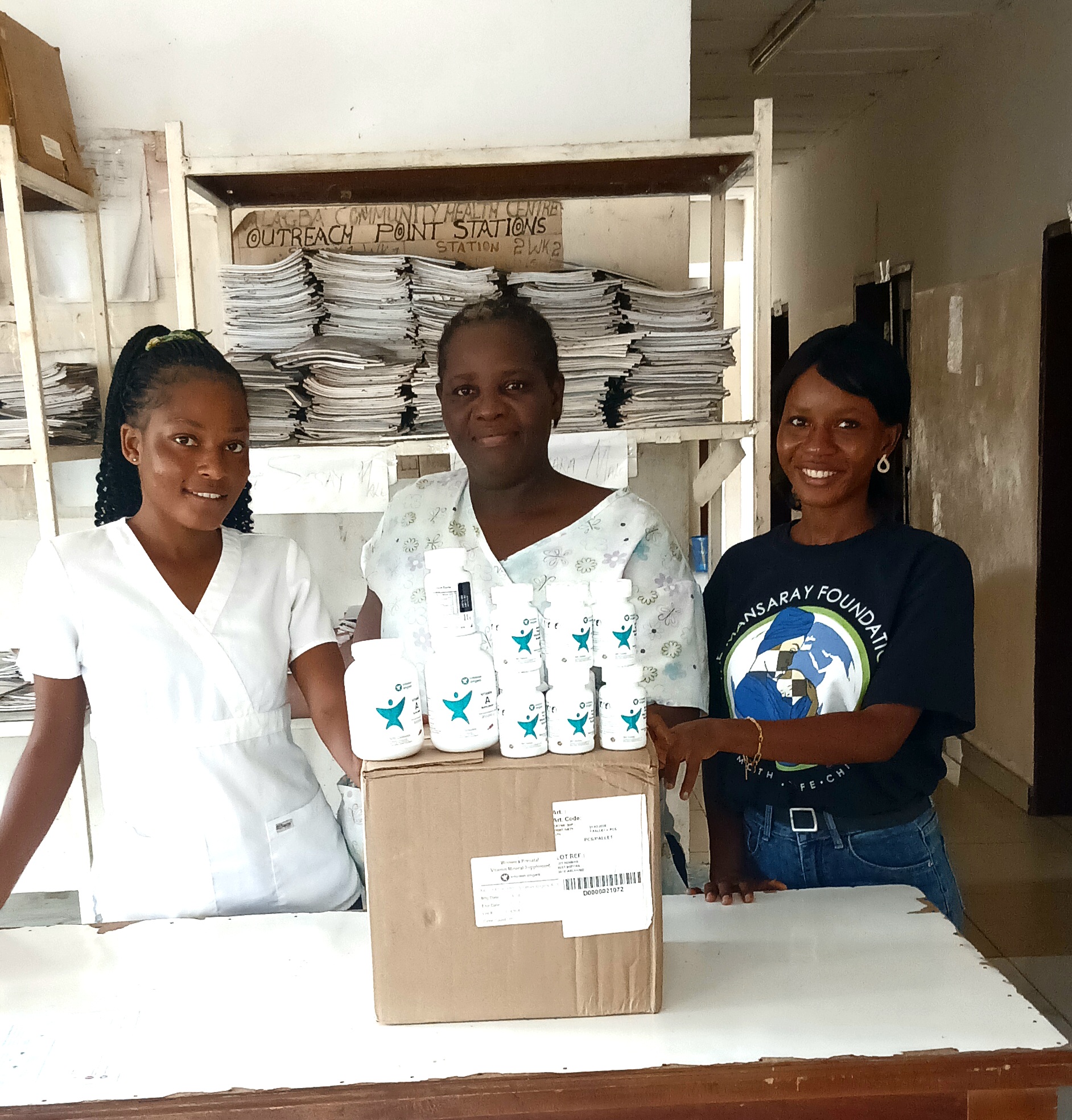 Two health workers and a TMF team member standing behind Vitamin Angels bottles and a box at a facility counter