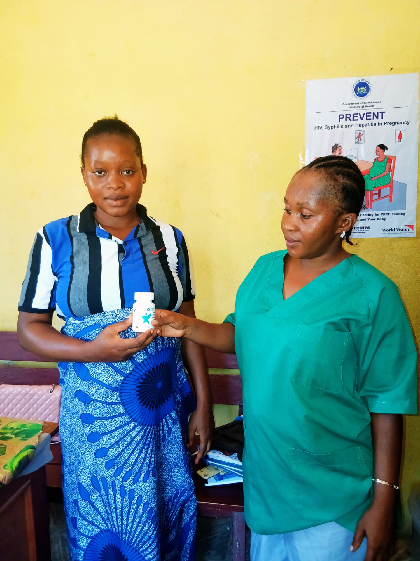 A health worker in green scrubs handing a Vitamin Angels bottle to a pregnant woman inside a health facility