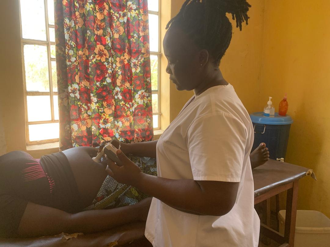 A health worker in white holding a handheld fetal Doppler to a pregnant woman's abdomen on an examination bed