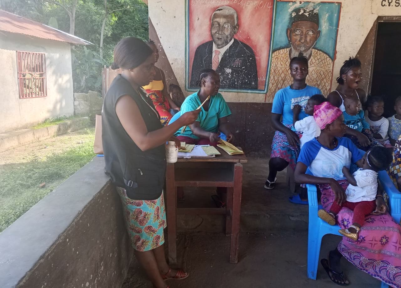 Women with young children seated on a veranda while a health worker and a TMF team member review cards at a table