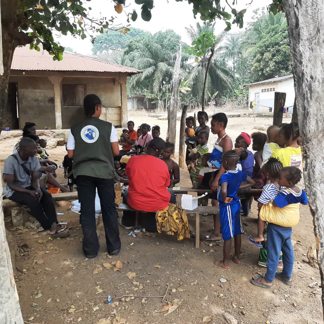 A TMF team member with women and children gathered on benches under trees in a community