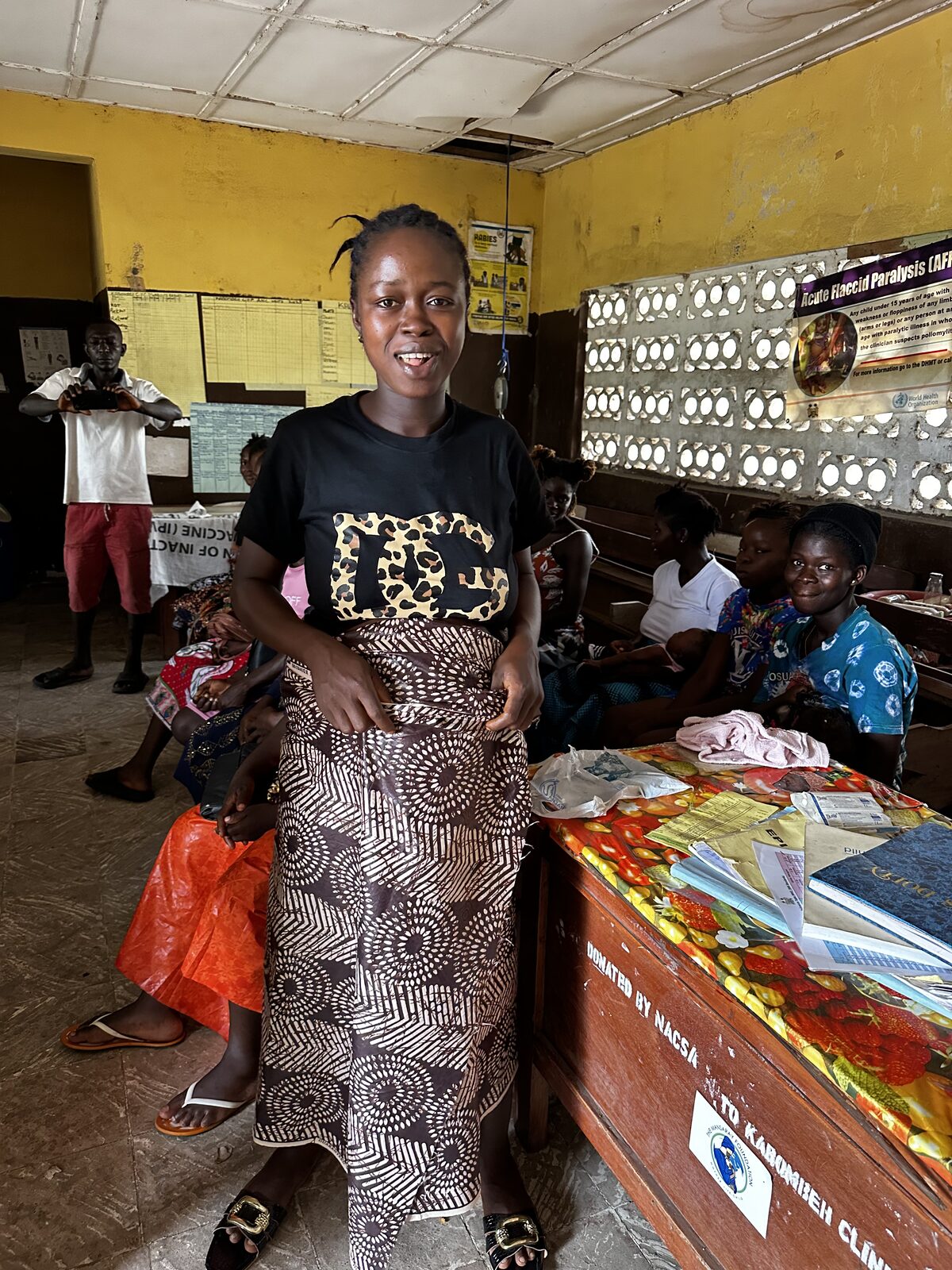 A smiling woman standing inside a clinic waiting area with other women seated on benches behind her
