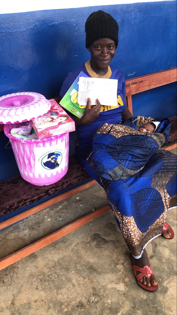 A mother seated with her baby beside a Safe Motherhood Birthing Bucket