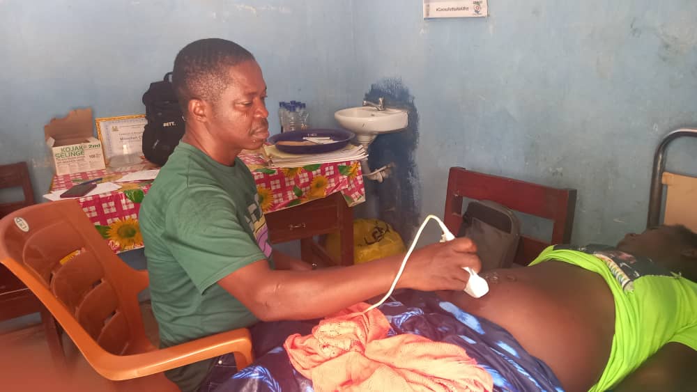 A health worker seated beside a patient, performing an abdominal ultrasound scan in a clinic room